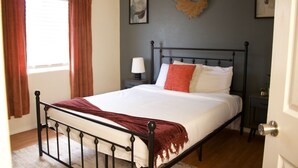 Una cama bien hecha con una colcha blanca y una manta roja, una mesita de noche con una lámpara, y una guirnalda en la pared.
