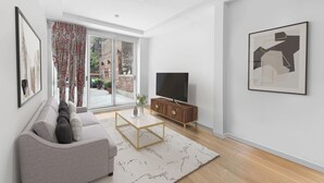 A modern living room with a grey sofa, a wooden cabinet, a flat-screen TV, and a large window with patterned curtains.