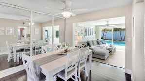 A modern dining area with a large table and white chairs, a ceiling fan, and a view of a pool and outdoor seating area. - Pet friendly house with a heated pool and hot tub! (Deerfield Beach)