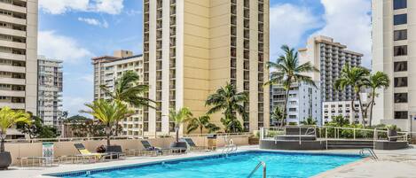 A rooftop pool area with lounge chairs and palm trees, surrounded by high-rise buildings.