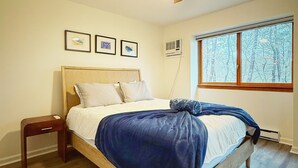 Ein Schlafzimmer mit einem Bett, einem Nachttisch, einem Deckenventilator, einem Fenster mit Blick auf Bäume und einem gemusterten Teppich.