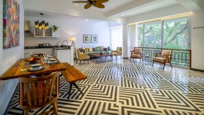 A spacious living area with a dining table, chairs, and a sofa. There is a kitchen in the background, a ceiling fan, and large windows with a view of trees. - El Rocio - 19 in Assagao (Assagao)