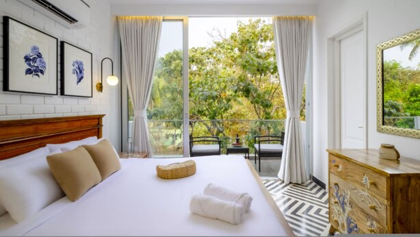 A bedroom with a large bed, a wooden dresser, a mirror, and a balcony with a view of greenery. - El Rocio - 19 in Assagao (Assagao)