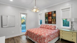 Une chambre à coucher comprenant un lit, une table de chevet, une fenêtre avec des stores, un luminaire au plafond et une porte donnant sur un balcon.