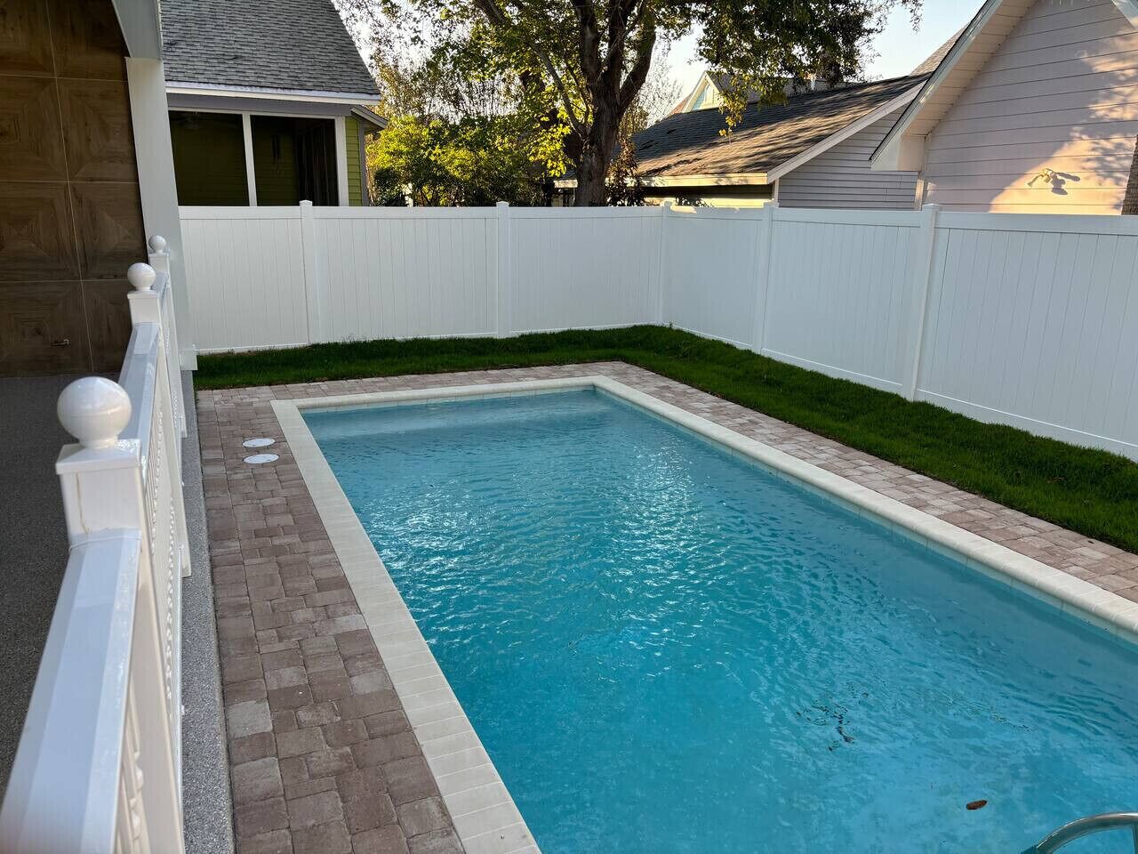 Une piscine rectangulaire bordée de briques, entourée d’une clôture blanche et d’une pelouse.