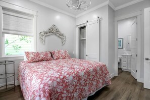 A bedroom with a floral bedspread, a chandelier, a wardrobe, and a door leading to another room.