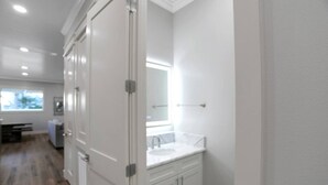 Un couloir avec une porte blanche, une salle de bain avec un lavabo et un miroir blancs, et une fenêtre donnant sur des arbres.