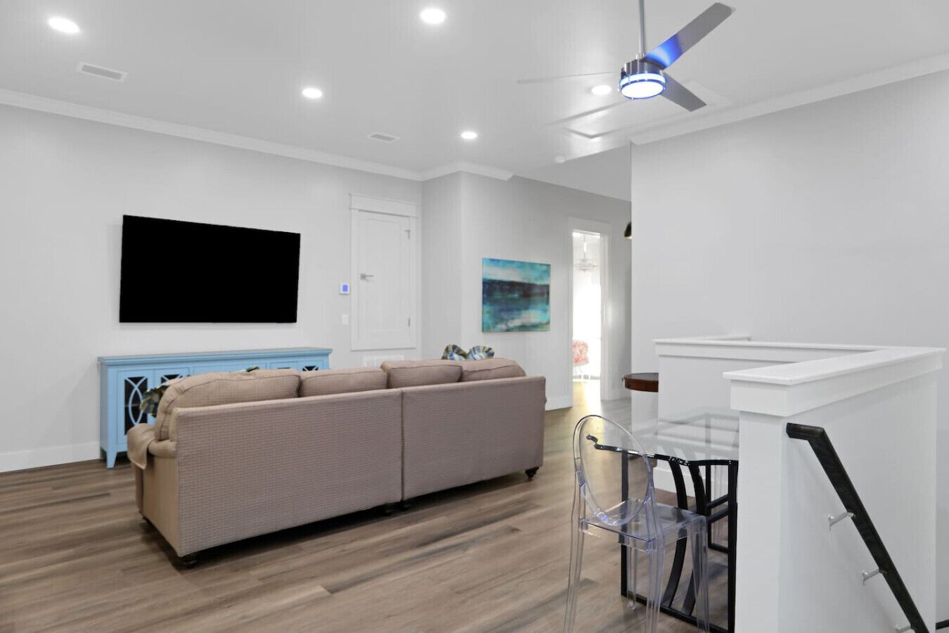 Un salon moderne avec un canapé d’angle beige, un téléviseur à écran plat fixé au mur, un ventilateur de plafond et un escalier doté d’une rampe en verre.