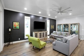 Un salon moderne avec un canapé gris, un fauteuil vert, un téléviseur à écran plat et un ventilateur de plafond.