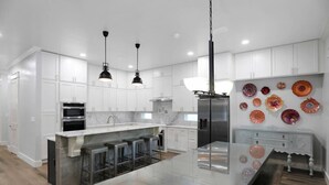 A modern kitchen with white cabinets, stainless steel appliances, a central island with stools, pendant lighting, and decorative glass plates on the wall. - Large Group Stay | Pool + Walk to Beach (Destin)