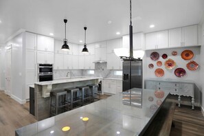 A modern kitchen with white cabinets, stainless steel appliances, a central island with stools, pendant lighting, and decorative glass plates on the wall. - Large Group Stay | Pool + Walk to Beach (Destin)