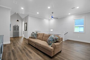Un salon spacieux avec un canapé beige, un plancher en bois, un ventilateur de plafond et une fenêtre équipée de stores.