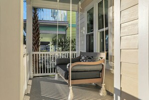 A porch with a swing chair, a white picket fence, and a view of a green building in the background.