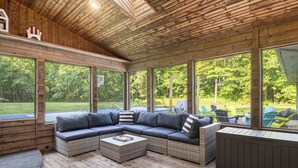 A covered outdoor seating area with a sectional sofa, wicker coffee table, and large windows offering views of a green landscape. - Secluded Retreat w/ Hot Tub, Pickleball/Basketball Court, Putting Green, Arcade (New Buffalo)