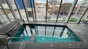 Piscina interior moderna com água cristalina, ladeada por azulejos pretos e paredes de vidro que oferecem vista para a cidade.