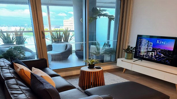 Uma sala de estar moderna com um sofá de couro, uma pequena mesa lateral de madeira e uma TV em um suporte branco.