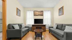 A living room with two grey sofas, a wooden coffee table, a TV on a wooden stand, and a fireplace. - Charming 1-Bedroom apartment in Sunnyside, Queens near 40th St-Lowery St station (Queens)