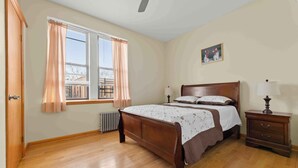 A bedroom with a wooden bed, two bedside tables with lamps, a radiator, and a window with curtains. - Charming 1-Bedroom apartment in Sunnyside, Queens near 40th St-Lowery St station (Queens)