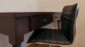 A black office chair with a cushioned seat and backrest, positioned in front of a dark wooden desk with a drawer.