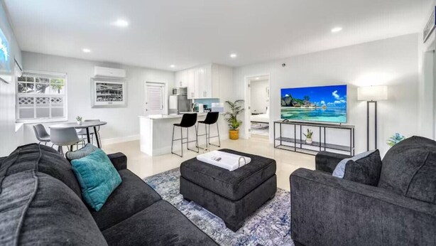 A modern living room with a grey sofa set, a coffee table, and a TV on a stand. There is a kitchen area with white cabinets and a dining table with chairs. - Coastal Unit .5 Miles to Beach! (Fort Lauderdale)