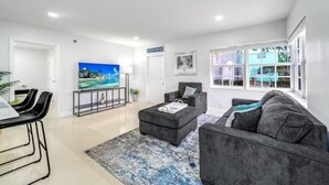 A modern living room with a flat-screen TV, grey sofas, a coffee table, and a dining area with black chairs. - Coastal Unit .5 Miles to Beach! (Fort Lauderdale)