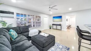 A modern living room with a grey sectional sofa, a small ottoman, a dining area with a table and chairs, and a large window with blinds. - Coastal Unit! .5 miles to Beach! (Fort Lauderdale)