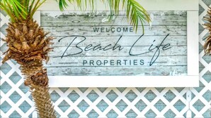 Ein Willkommen-Schild mit dem Aufdruck "Welcome Beach Life Properties" vor einem Holzhintergrund, eingerahmt von Palmen.
