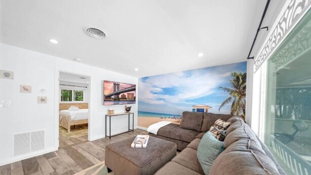 Ein Wohnzimmer mit einer großen Eckcouch, einem Couchtisch und einem Fernseher. An der Wand ist eine Strandansicht zu sehen und aus dem Fenster blickt man auf Palmen.