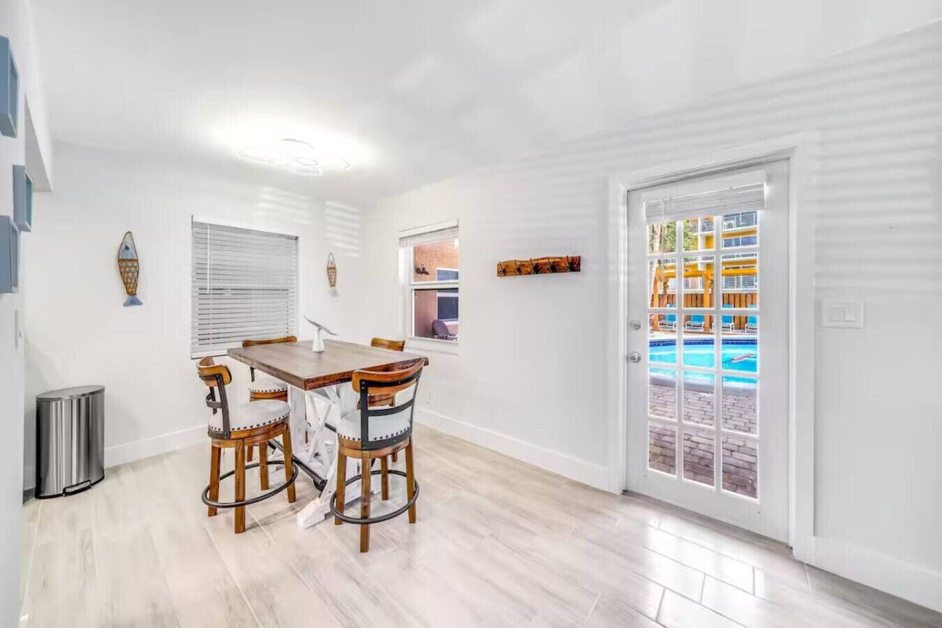A dining area with a wooden table and chairs, a trash can, and a door leading to a pool area.