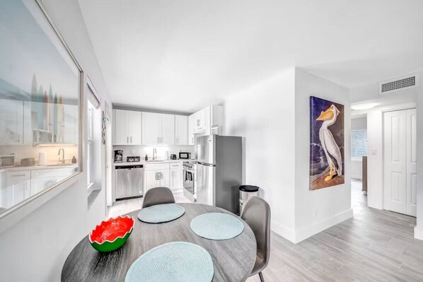 A modern kitchen with white cabinets, a microwave, and a refrigerator. There is a dining area with a table and chairs, and a large painting on the wall.