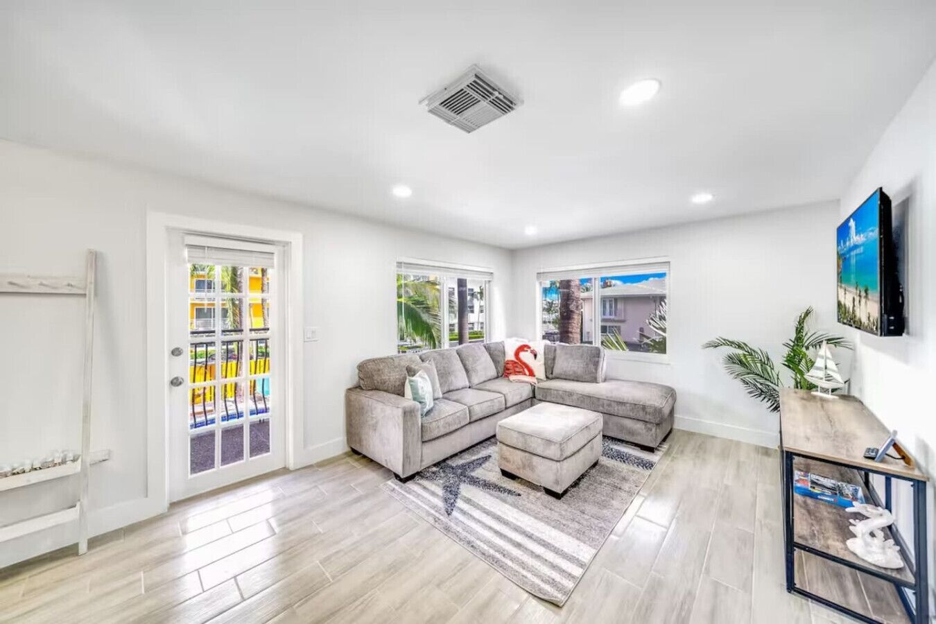 A modern living room with a grey sectional sofa, a small ottoman, and a striped rug. There are two windows with views of palm trees and a building.