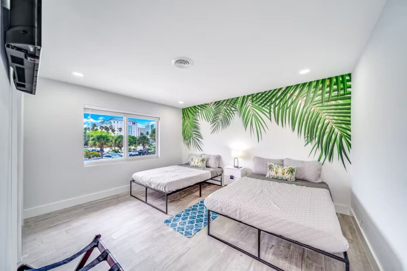 Un dormitorio moderno con dos camas, un ventanal con vistas a la ciudad y una pared decorada con hojas de palmera verdes.