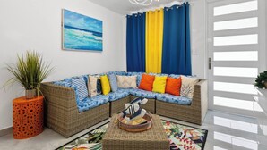 Una sala de estar con un sofá de mimbre, cojines coloridos, una planta en maceta y un cuadro de una playa.