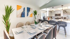A modern dining area with a white table set for six, surrounded by chairs. There are abstract paintings on the wall, and a potted plant on the left.