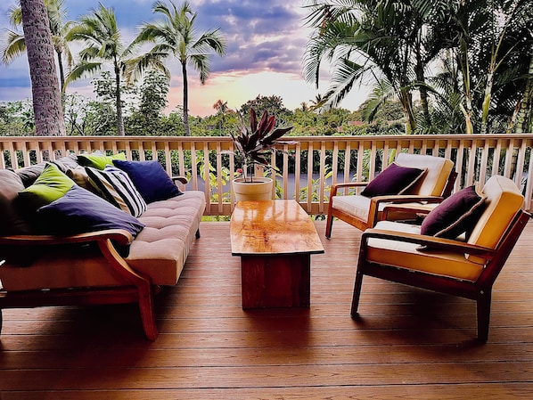 A wooden deck with a sofa and two chairs, a table, and a view of palm trees and a sunset. - NEW 1-bedroom bungalow with AC, WiFi in marvelous Kailua-Kona Aug specials (Kailua-Kona)