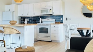 Eine moderne Küche mit weißen Schränken, einer Mikrowelle und einem Backofen. Zwei Korbstühle und ein runder Holztisch mit einem Buch sind ebenfalls vorhanden. Die Decke ist mit Holzbalken versehen und es gibt zwei Kronleuchter.