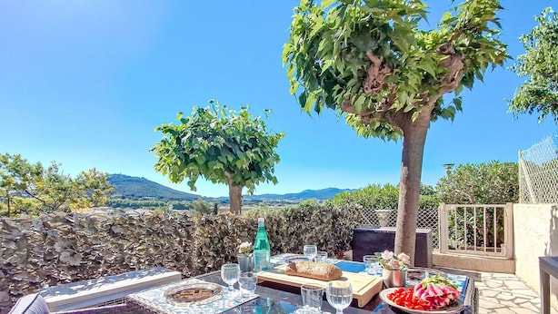 Un espace extérieur aménagé pour les repas, avec une table dressée pour deux personnes, entouré de meubles en osier, offrant une vue sur la campagne et bordé d’un mur en pierre.