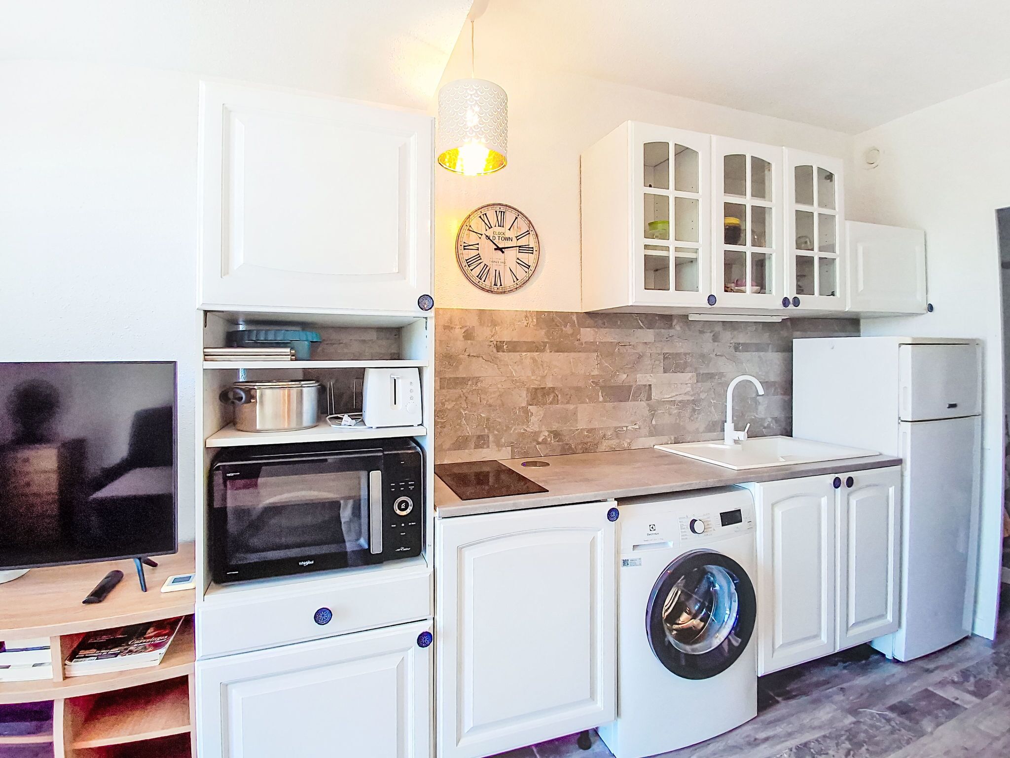 Una cocina compacta con armarios blancos, microondas, lavadora, pileta y un reloj en la pared.