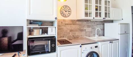 Una cocina compacta con armarios blancos, microondas, lavadora, pileta y un reloj en la pared.