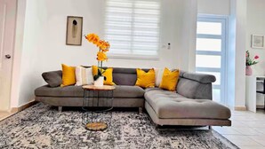A modern living room with a grey sectional sofa, a round coffee table, and a large window with blinds. - La Casa de Ada Near Airport & Beaches Charming 3-bedroom house with AC andWiFi (San Juan)