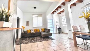 A spacious living room with a grey sectional sofa, a coffee table, and a dining area with a glass table and chairs. - La Casa de Ada Near Airport & Beaches Charming 3-bedroom house with AC andWiFi (San Juan)