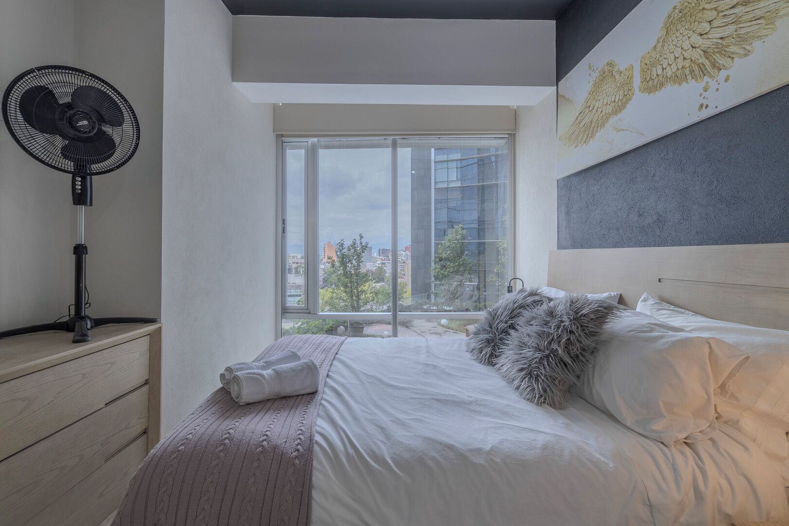 Un dormitorio con cama, un ventilador de pie, una ventana con vista a la ciudad y una pared decorada con una obra de arte de plumas.