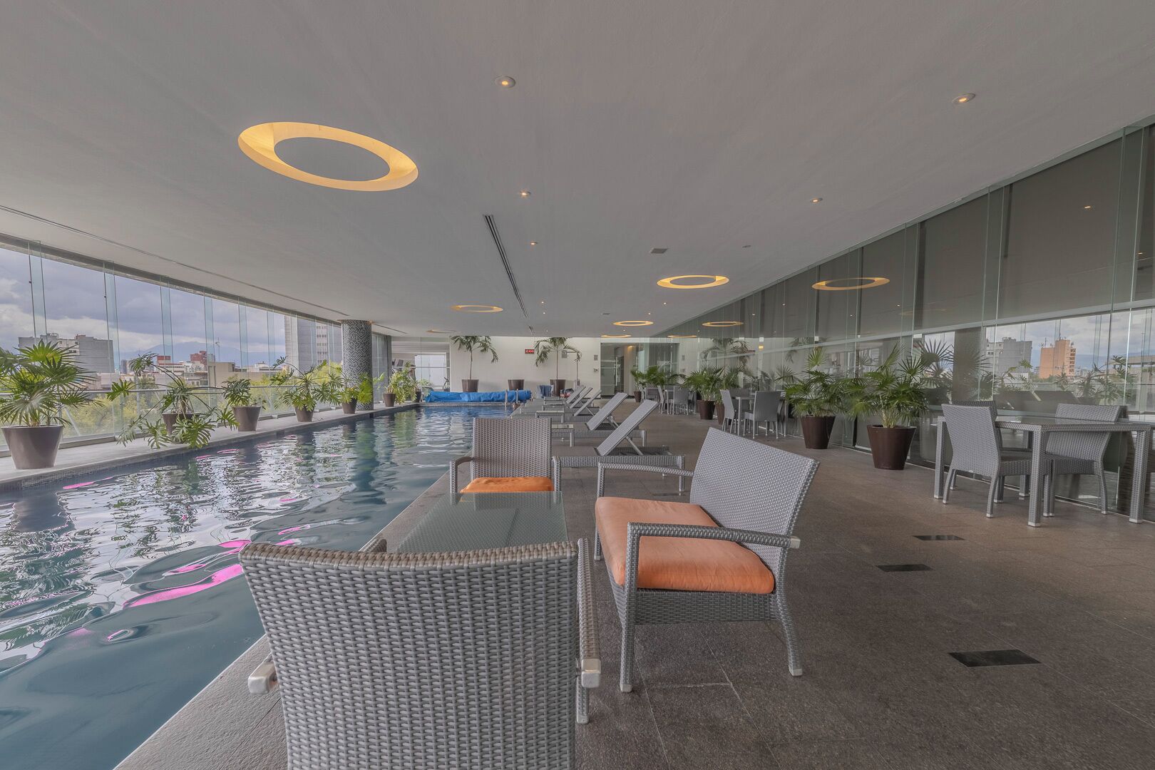 Un área de piscina interior moderna con sillones y mesas, rodeada de amplios ventanales con vistas a la ciudad.