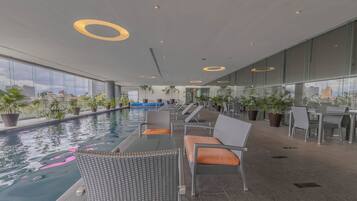 Un área de piscina interior moderna con sillones y mesas, rodeada de amplios ventanales con vistas a la ciudad.