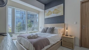 A bedroom with a large window, a bed with rolled towels, a nightstand with a lamp, and a wooden door. - Apt with Reforma View - Pool and Jacuzzi (Mexico City)