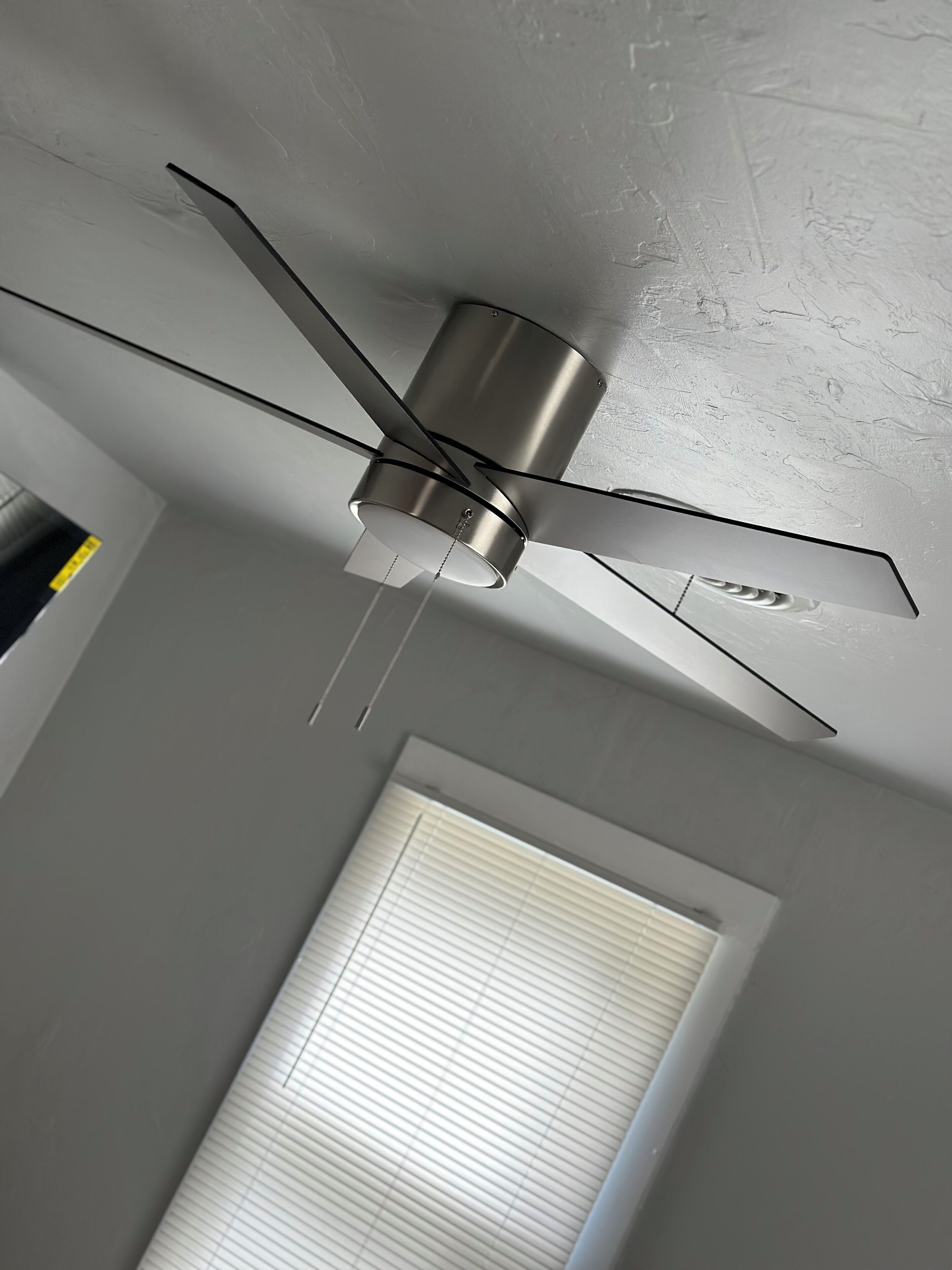 A ceiling fan with a metallic finish, a window with blinds, and a textured ceiling.