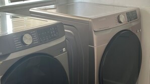 A front-loading Samsung washer and dryer in a laundry room with a window and blinds.