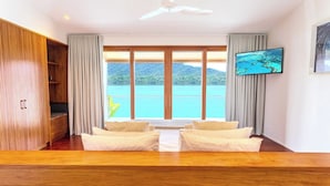 Um quarto com piso de madeira, dois sofás brancos, um ventilador de teto, uma TV na parede e uma janela grande com vista para o mar e montanhas.