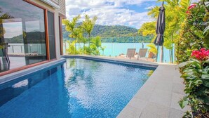 Piscina com vista para um lago e montanhas, cercada por plantas tropicais e móveis de exterior.