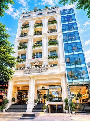 Un immeuble à plusieurs étages doté de balcons, une enseigne indiquant « GOLDEN SUN 7 APARTMENTS », et un bâtiment moderne en verre situé à droite.